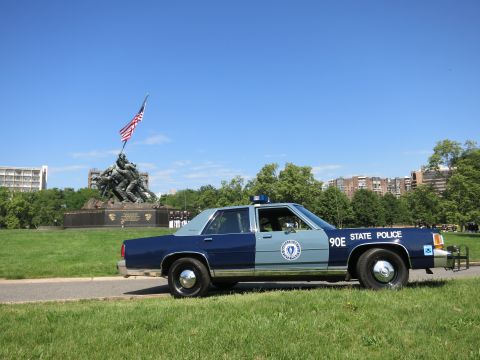 1991 Ford LTD Crown Victoria - blue Massachusetts State Police sedan marked 90E parked on grass with the Iwo Jima Memorial and city buildings in the distance.