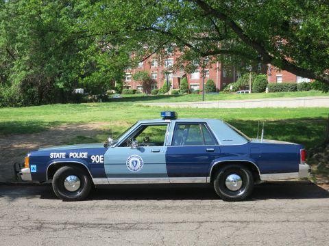 1991 Ford LTD Crown Victoria - Driver-side profile of a two-tone blue Massachusetts State Police sedan marked 90E parked on pavement near brick buildings.