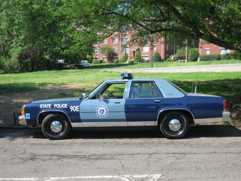 1991 Ford LTD Crown Victoria - Driver-side profile of a blue and light-blue Massachusetts State Police sedan marked 90E parked along a curb with a brick building behind.