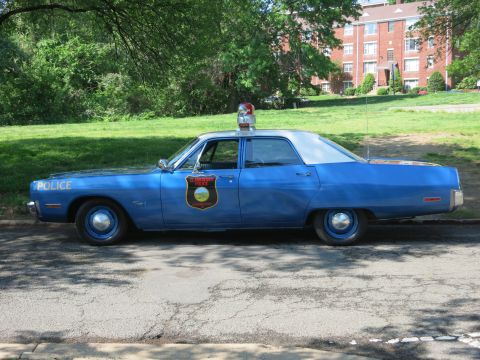 1991 Ford LTD Crown Victoria - Side view of a weathered light-blue vintage police sedan with a single rooftop beacon and a gold shield door emblem, parked on pavement.