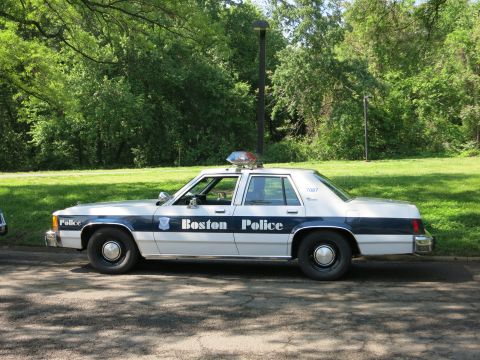 1991 Ford LTD Crown Victoria - Side view of a blue and white sedan lettered Boston Police with a rooftop beacon, parked along a curb by trees.