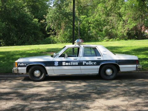1991 Ford LTD Crown Victoria - Driver-side profile of a blue and white Boston Police sedan with a rooftop beacon parked along a shaded curb.