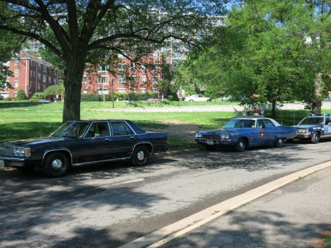 1991 Ford LTD Crown Victoria - dark sedan and two marked police cruisers parked in a row along a curb with brick buildings and lawn behind.
