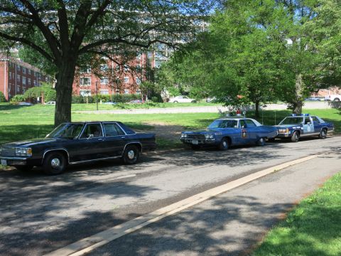 1991 Ford LTD Crown Victoria - dark sedan beside two marked vintage police cruisers parked along a shaded curb with brick buildings in the background.