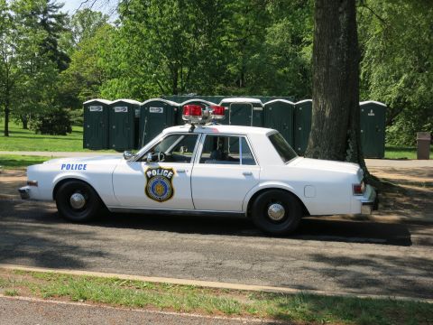 1991 Ford LTD Crown Victoria - Side view of a white police sedan with a rooftop light bar and gold shield emblem parked near a row of portable toilets.
