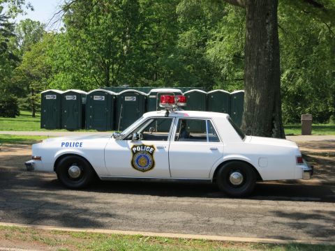 1991 Ford LTD Crown Victoria - Driver-side profile of a white police sedan with a rooftop light bar and shield door emblem parked before green portable toilets.