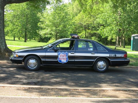 1991 Ford LTD Crown Victoria - Side view of a black State Patrol sedan with a blue-and-white door shield and State Trooper lettering, parked on a shaded street.
