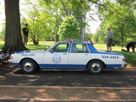 1991 Ford LTD Crown Victoria - Side view of a blue and white police sedan lettered Police and 334-2424 with a rooftop light bar, with people standing nearby on grass.