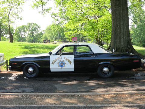 1991 Ford LTD Crown Victoria - Driver-side profile of a black and white Highway Patrol sedan lettered with a gold star emblem and rooftop beacon, parked under a tree.