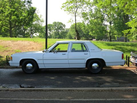 1991 Ford LTD Crown Victoria - white Ford Crown Victoria sedan sits in left profile on a roadway beside a grassy park with trees.