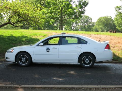 1991 Ford LTD Crown Victoria - white Chevrolet Impala police sedan with a roof light bar and door shield decal is parked in left profile by a park.