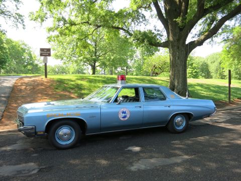1991 Ford LTD Crown Victoria - light blue full-size Buick police sedan with a red dome light and 'Police' lettering parked under trees in a park.