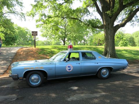 1991 Ford LTD Crown Victoria - light blue full-size Buick police sedan with red dome light and door shield parked in profile beside a shaded park road.