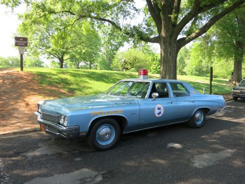 1991 Ford LTD Crown Victoria - light blue Buick police sedan with red dome light leads a row of parked vintage police cars along a tree-lined park road.