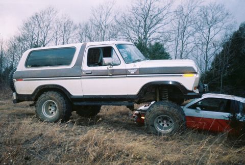 1978 Ford Bronco - A lifted 1978 Ford Bronco is positioned over a crushed car in a grassy, wooded area.