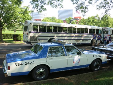 1991 Ford LTD Crown Victoria - line of cars, one with its hood up, parked along a road below a grassy embankment and elevated highway under blue sky.