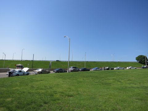 1991 Ford LTD Crown Victoria - row of cars parked along a roadway beside a grassy embankment with an elevated overpass and streetlights under blue sky.
