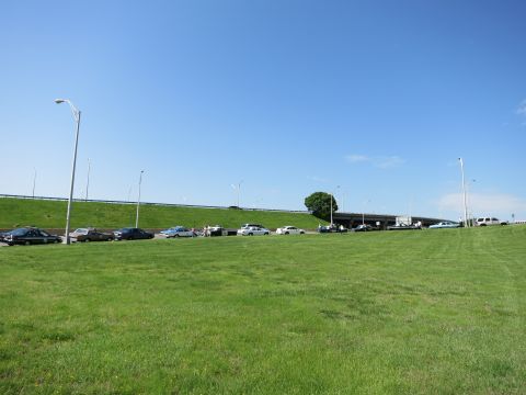 1991 Ford LTD Crown Victoria - Several cars parked along a roadway below an elevated highway bridge, with a broad grassy field under bright blue sky.