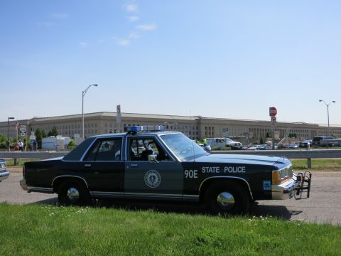 1991 Ford LTD Crown Victoria - black and gray State Police Ford sedan marked 90E with roof lights, parked on grass in front of the Pentagon building.