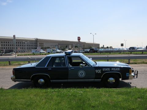 1991 Ford LTD Crown Victoria - Full side profile of a black and gray State Police Ford sedan marked 90E parked on a roadside before the Pentagon.