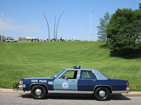 1991 Ford LTD Crown Victoria - blue and light-blue State Police Ford sedan marked 90E parked on a road with the Air Force Memorial spires on the green hill behind.