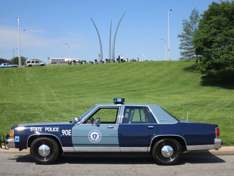 1991 Ford LTD Crown Victoria - two-tone blue State Police Ford sedan marked 90E with door seal, parked below the Air Force Memorial spires on a grassy hill.