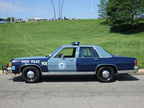 1991 Ford LTD Crown Victoria - two-tone blue State Police Ford sedan marked 90E with door seal parked on a road below a grassy hill.
