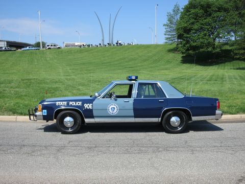 1991 Ford LTD Crown Victoria - two-tone blue State Police Ford sedan numbered 90E parked roadside with the Air Force Memorial spires on the hill behind.