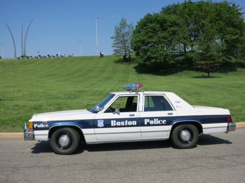 1991 Ford LTD Crown Victoria - Profile of a white and blue 'Boston Police' Ford LTD sedan with roof lights parked on a road by a grassy hill and trees.