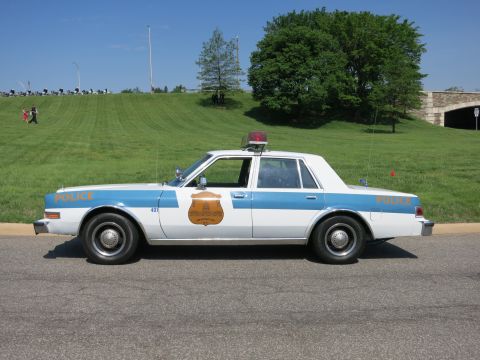 1991 Ford LTD Crown Victoria - light blue and white 'Police' sedan numbered 401 with gold door emblem and roof lights parked on a road by a grassy hill.
