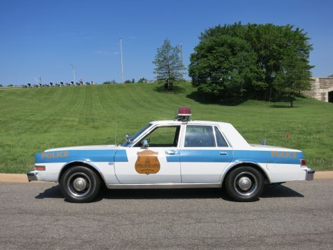 1991 Ford LTD Crown Victoria - Profile of a light blue and white 'Police' sedan marked 401 with gold door seal and red roof light parked on a road.