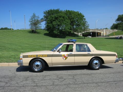1991 Ford LTD Crown Victoria - Profile of a tan and brown 'State Police' Chevrolet sedan with Maryland-style shield emblem and roof light parked on a road.