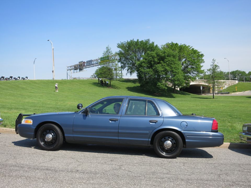 Dark blue Ford Crown Victoria sedan in profile with steel wheels, parked on a road by a hill
