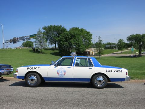 1991 Ford LTD Crown Victoria - white-and-blue police sedan marked POLICE and the number 334-2424 with a roof beacon is parked on a road by a grassy hill.