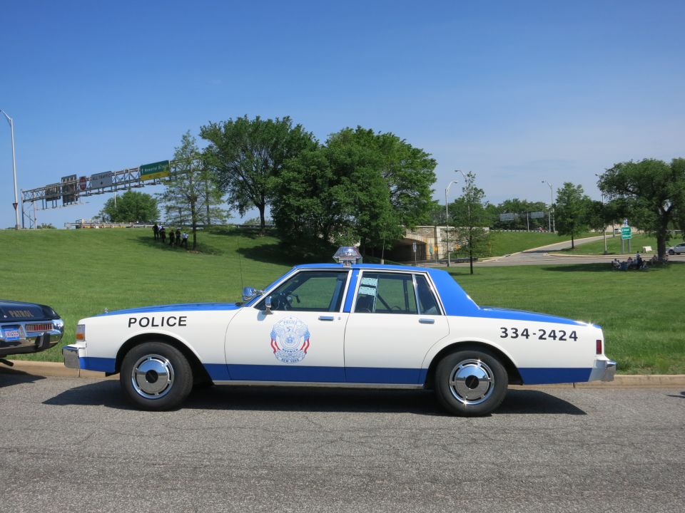 White and blue police sedan reading POLICE and 334-2424 in profile, parked roadside