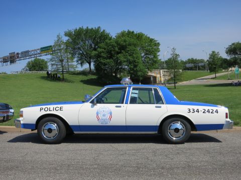 1991 Ford LTD Crown Victoria - white-and-blue police sedan lettered POLICE and 334-2424 with a roof light bar sits parked beside a green hillside.