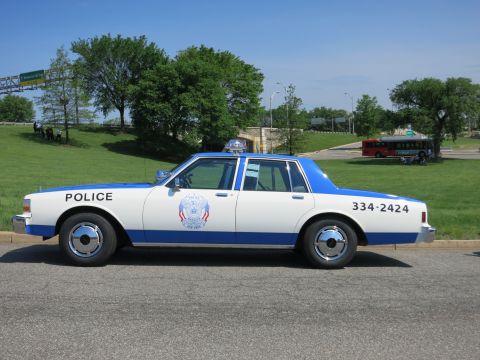 1991 Ford LTD Crown Victoria - white-and-blue police sedan marked POLICE and 334-2424 is parked on a road with a red bus visible in the distance.