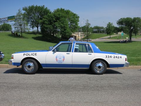 1991 Ford LTD Crown Victoria - white-and-blue police cruiser lettered POLICE and 334-2424 with a roof beacon sits parked beside a green slope.