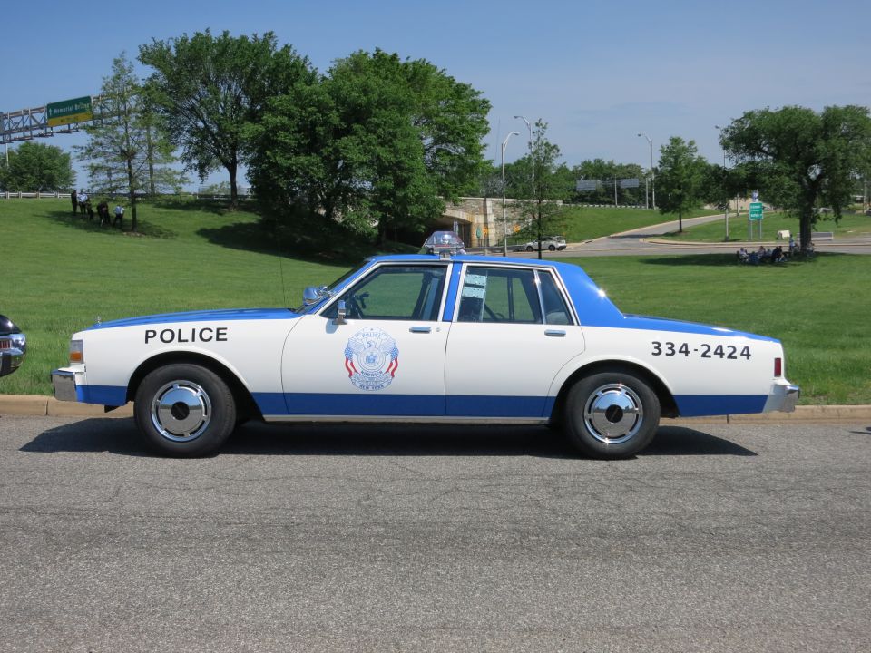 White and blue POLICE cruiser 334-2424 with door shield, parked by a grassy hill