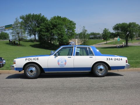 1991 Ford LTD Crown Victoria - white-and-blue police sedan marked POLICE and 334-2424 with a roof light bar is parked roadside under clear blue sky.