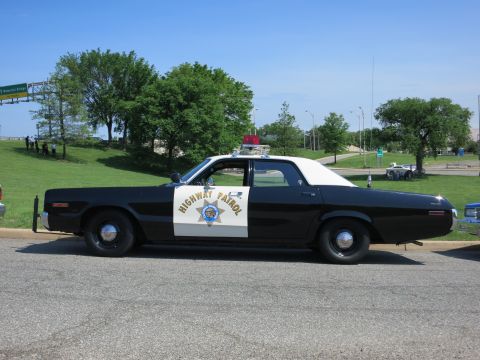 1991 Ford LTD Crown Victoria - black-and-white Highway Patrol sedan with a red roof light and gold star door shield sits parked on a road by a grassy hill.