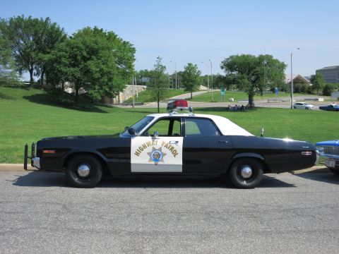 1991 Ford LTD Crown Victoria - black-and-white Highway Patrol sedan marked with a gold star and roof beacon is parked roadside beside a green hill.