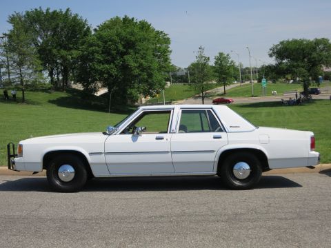 1991 Ford LTD Crown Victoria - white Ford LTD Crown Victoria sedan with a front push bar and black steel wheels is parked on a road by a grassy field.