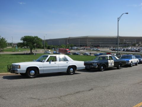 1991 Ford LTD Crown Victoria - white sedan leads a line of vintage police cruisers parked along a road in front of the Pentagon building.