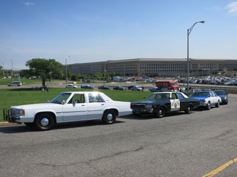 1991 Ford LTD Crown Victoria - white police sedan and a row of vintage cruisers are parked along a road in front of the Pentagon under clear sky.