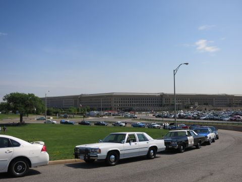 1991 Ford LTD Crown Victoria - White and other police cruisers are parked in a row along a road in front of the Pentagon and a busy parking lot.