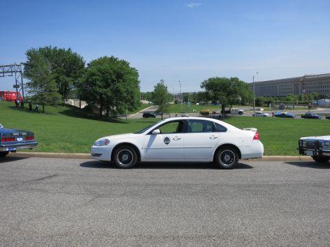 1991 Ford LTD Crown Victoria - white modern Chevrolet Impala police sedan with a door emblem is parked roadside near the Pentagon and a grassy slope.