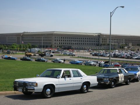 1991 Ford LTD Crown Victoria - white sedan leads a line of vintage and modern police cars parked along a road in front of the Pentagon.