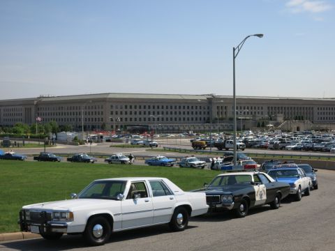 1991 Ford LTD Crown Victoria - white sedan and a line of police cruisers are parked along a road in front of the Pentagon under bright blue sky.