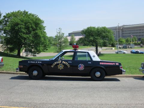 1991 Ford LTD Crown Victoria - black-and-white Highway Patrol sedan with a red roof light, To Protect & Serve and EMERGENCY 911 lettering is parked by a hill.
